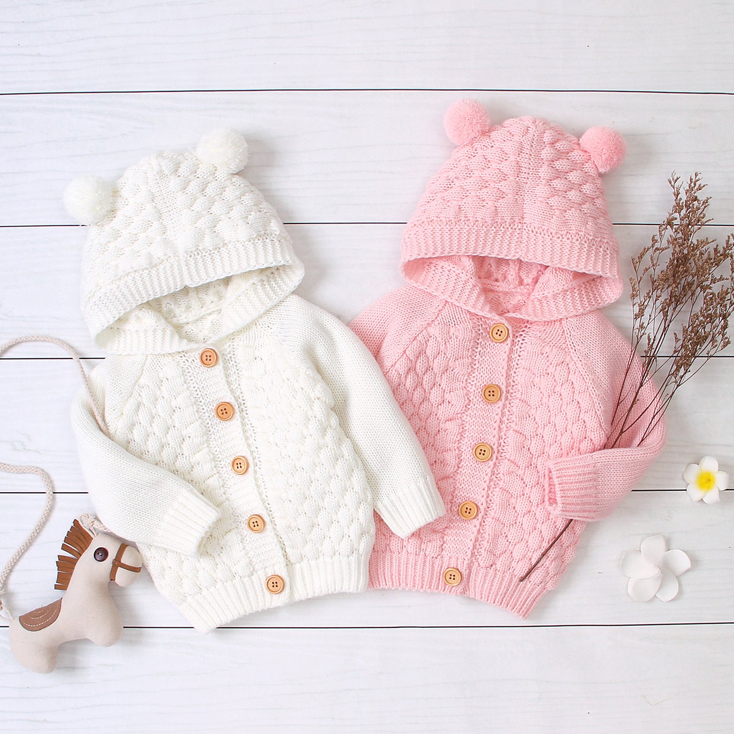 White and pink knitted baby cardigans with hats on a light wooden surface.