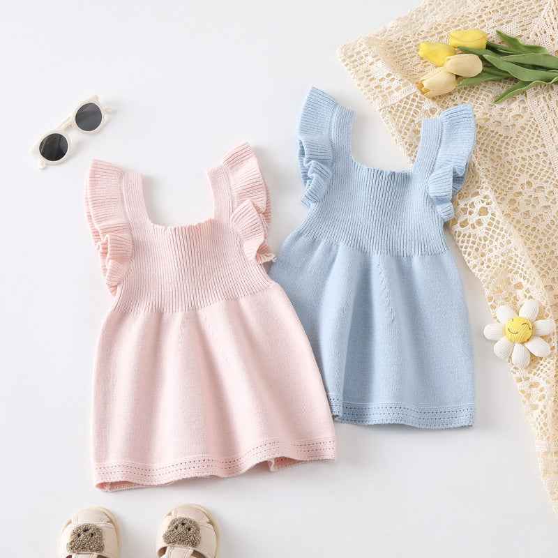 Two baby dresses, one pink and one blue, on a light surface with flowers and sunglasses.