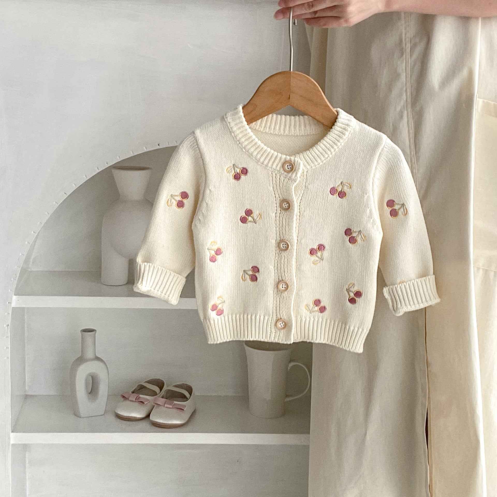White children's cardigan with cherry embroidery on a wooden hanger against a neutral background.